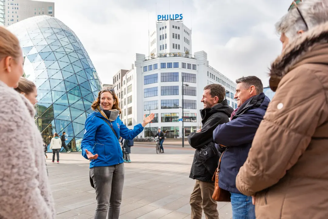 A guide talking to guests on the square by the glass Blob and the Philips Lichttoren, Eindhoven