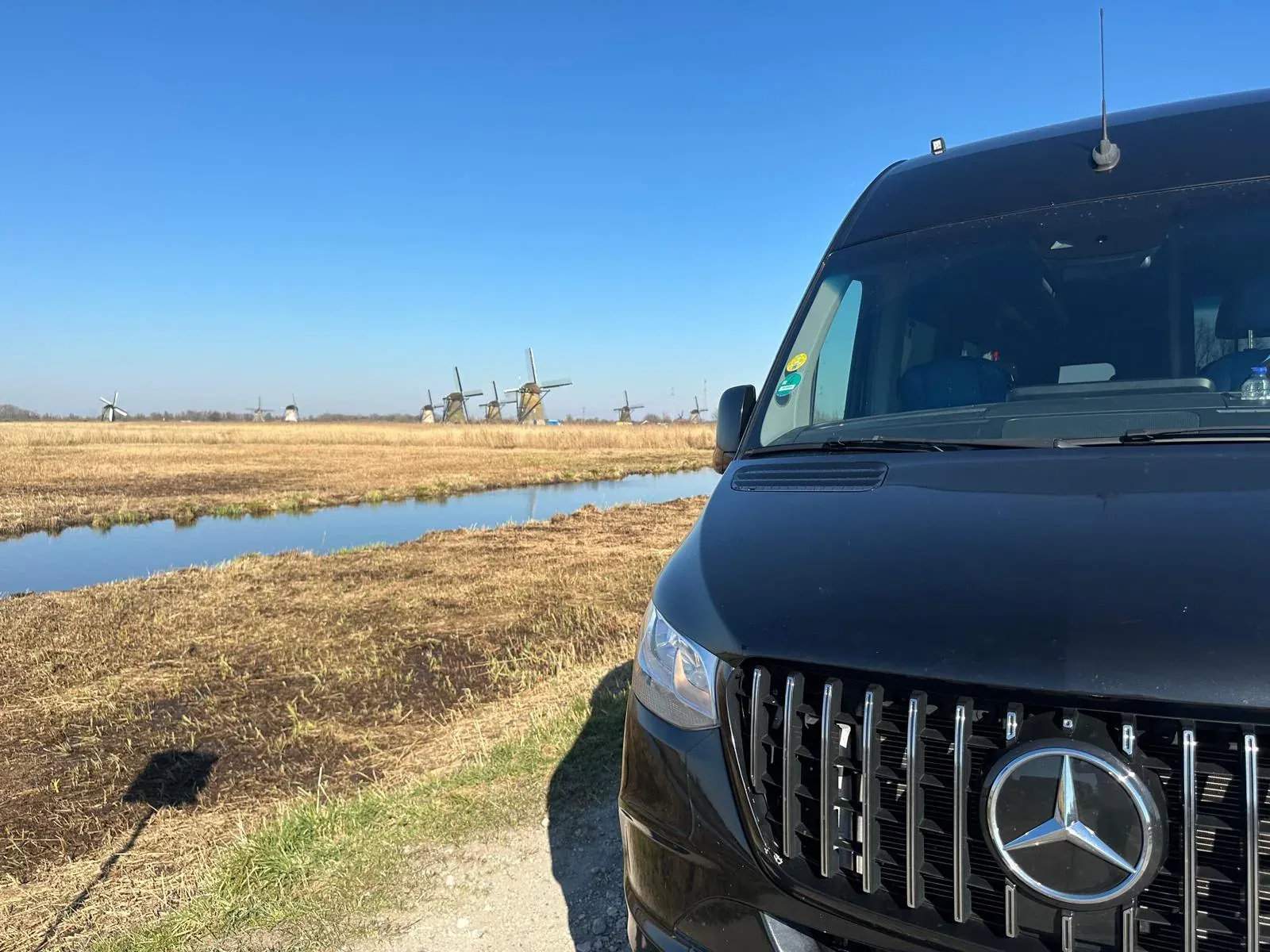 The Holland City Tours minivan parked beside a polder ditch at Kinderdijk, with the windmills lined up across the field behind it